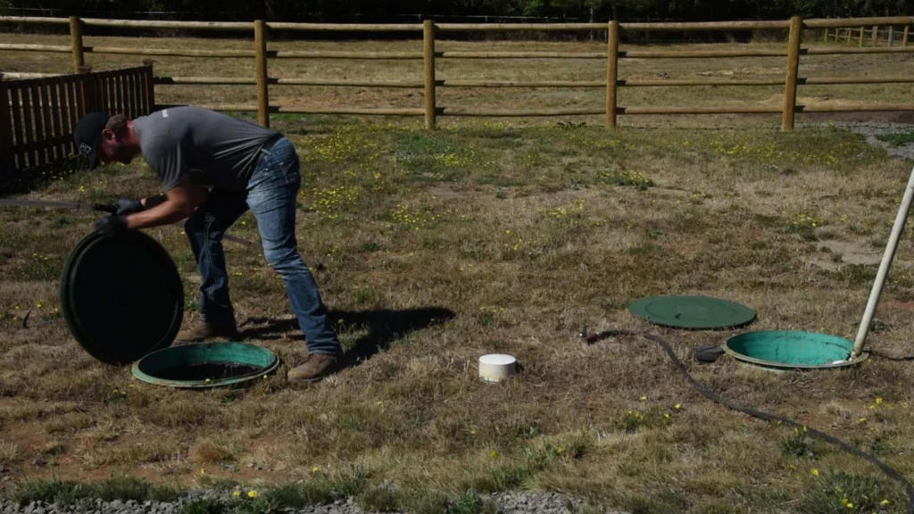 A technician lifting the green lid off a septic tank riser in a fenced yard.