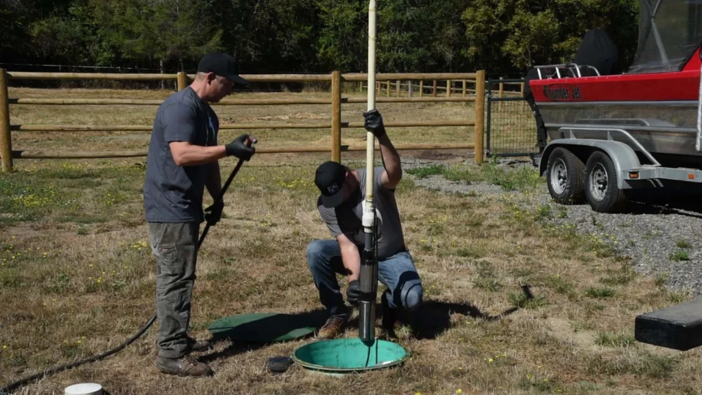 Two technicians lowering a sludge probe into an open septic tank.