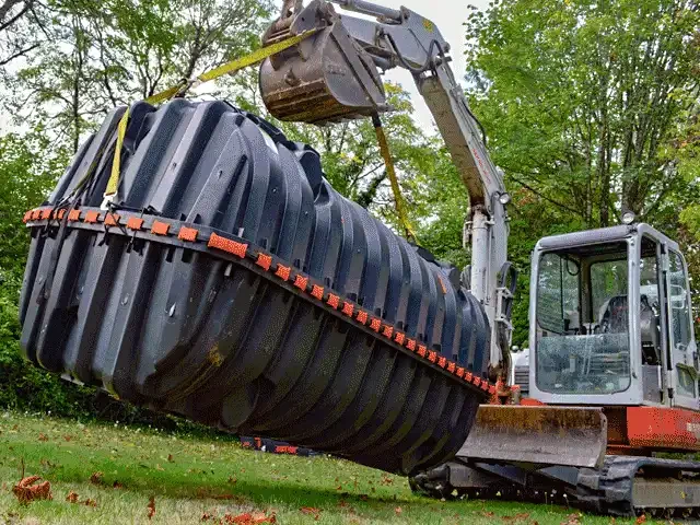 An excavator lowering a new black polyethylene septic tank into place.
