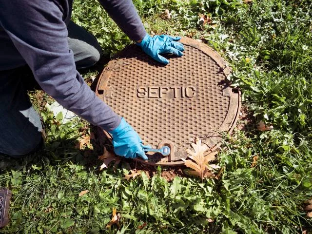 A gloved technician lifting a cast iron septic lid in the grass.